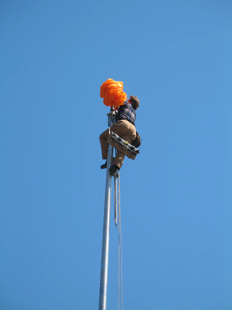 Climber dressing a Nishan Sahib flagpole in saffron cloth at a Sikh gurdwara
