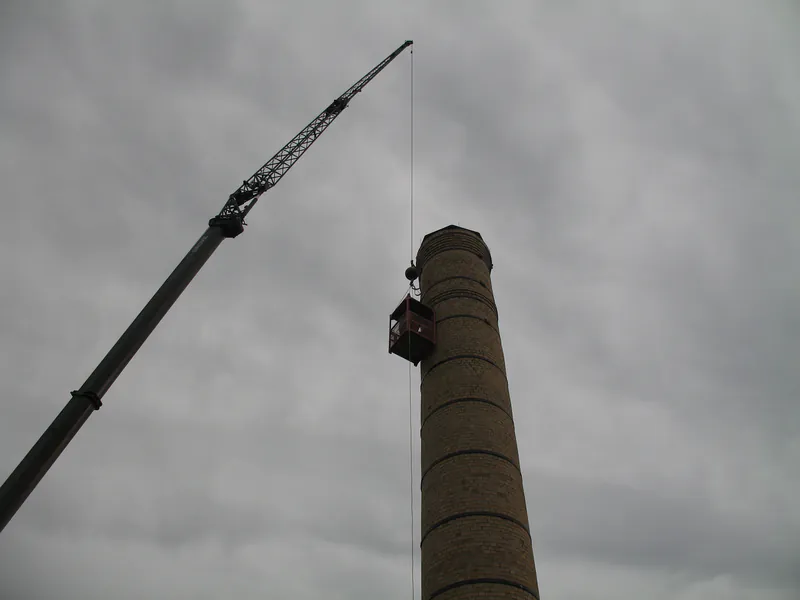 Man basket on a lattice crane alongside a brick smokestack